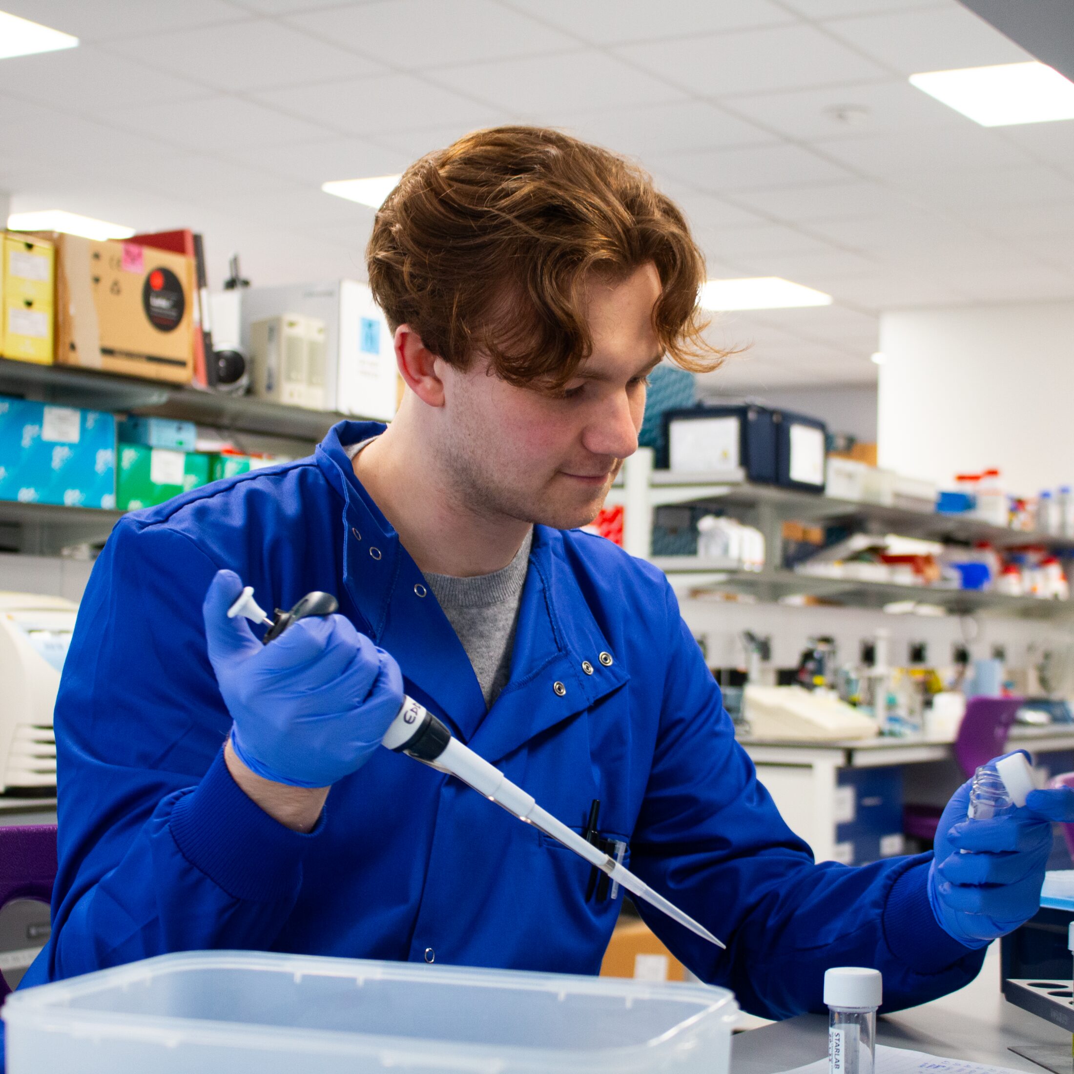 A person wearing a blue lab coat and gloves uses a micropipette to transfer liquid into a small tube in a laboratory setting.