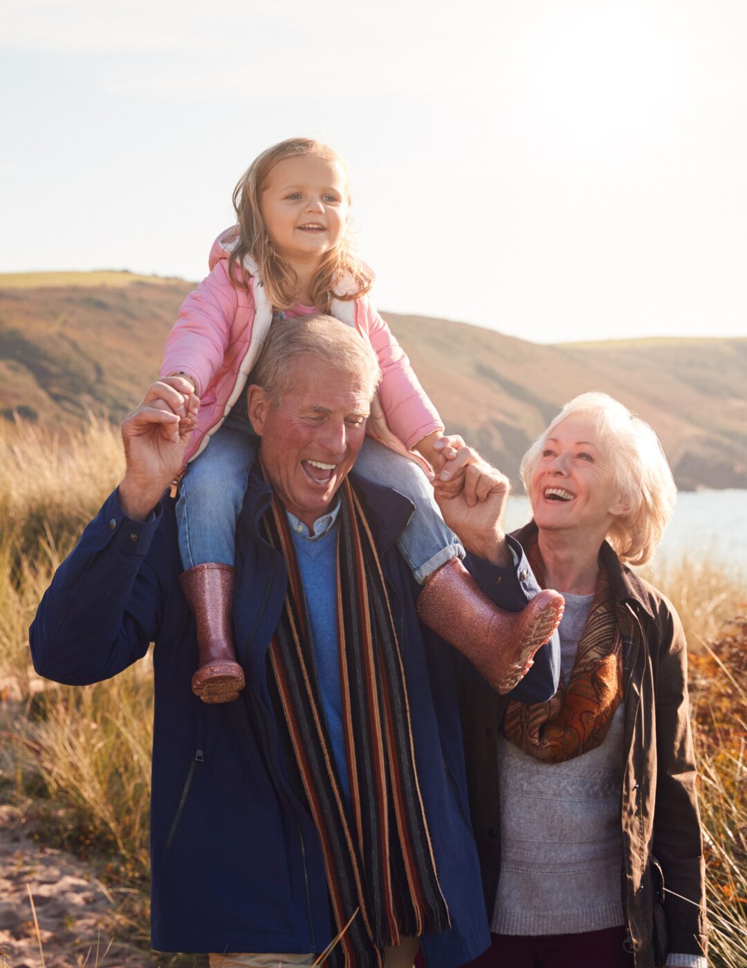 A group of three people walk outdoors through grassy landscape near a coastline, with a small child sitting on the shoulders of one of the adults.