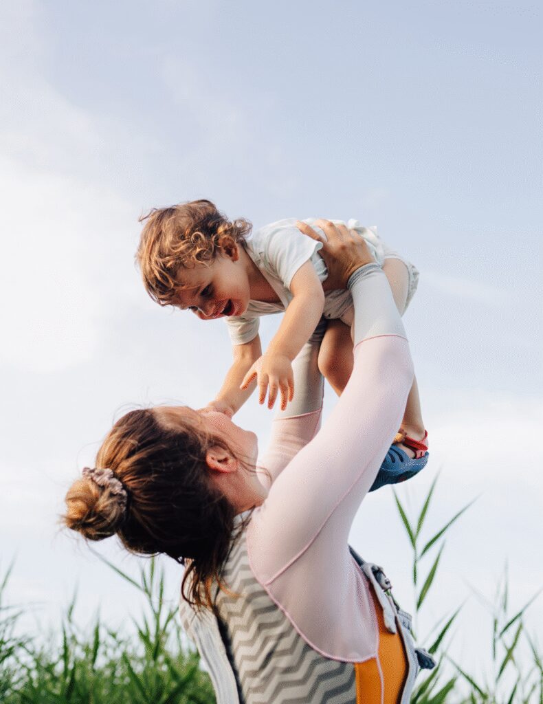 An adult lifts a small child into the air outdoors, with tall green plants and a bright sky in the background.
