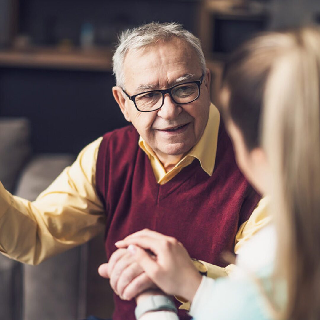 One person wearing a long‑sleeved shirt and a sleeveless sweater gestures with one hand while another person sits nearby holding the first person’s other hand in a home-like setting.