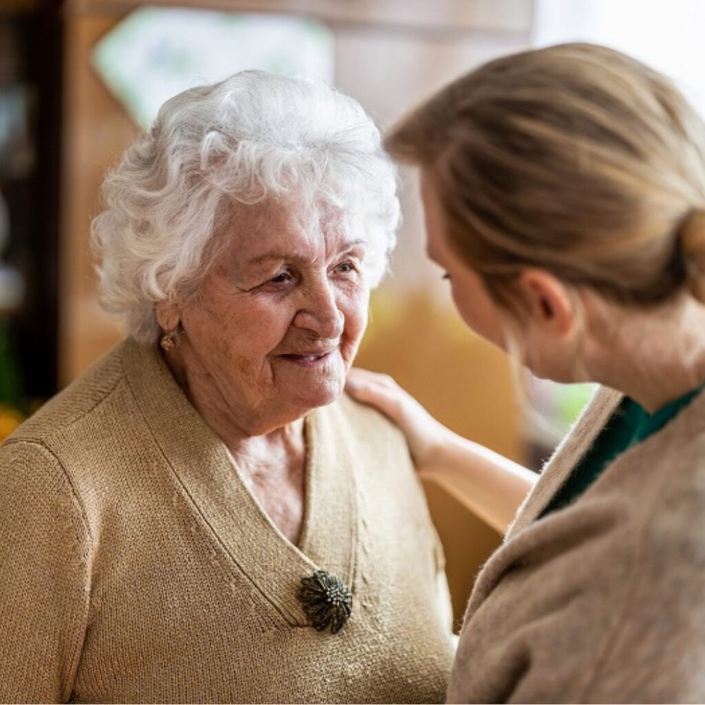 One person wearing a light-colored sweater places a hand on the shoulder of an older person with white hair who is standing nearby in an indoor setting.